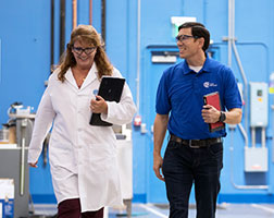 Image de deux personnes qui marchent dans une salle de laboratoire avec des planchettes à pince à la main.